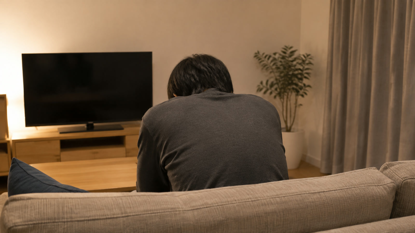 夜のリビングで、ソファに座って肩を落としている人の後ろ姿の写真
